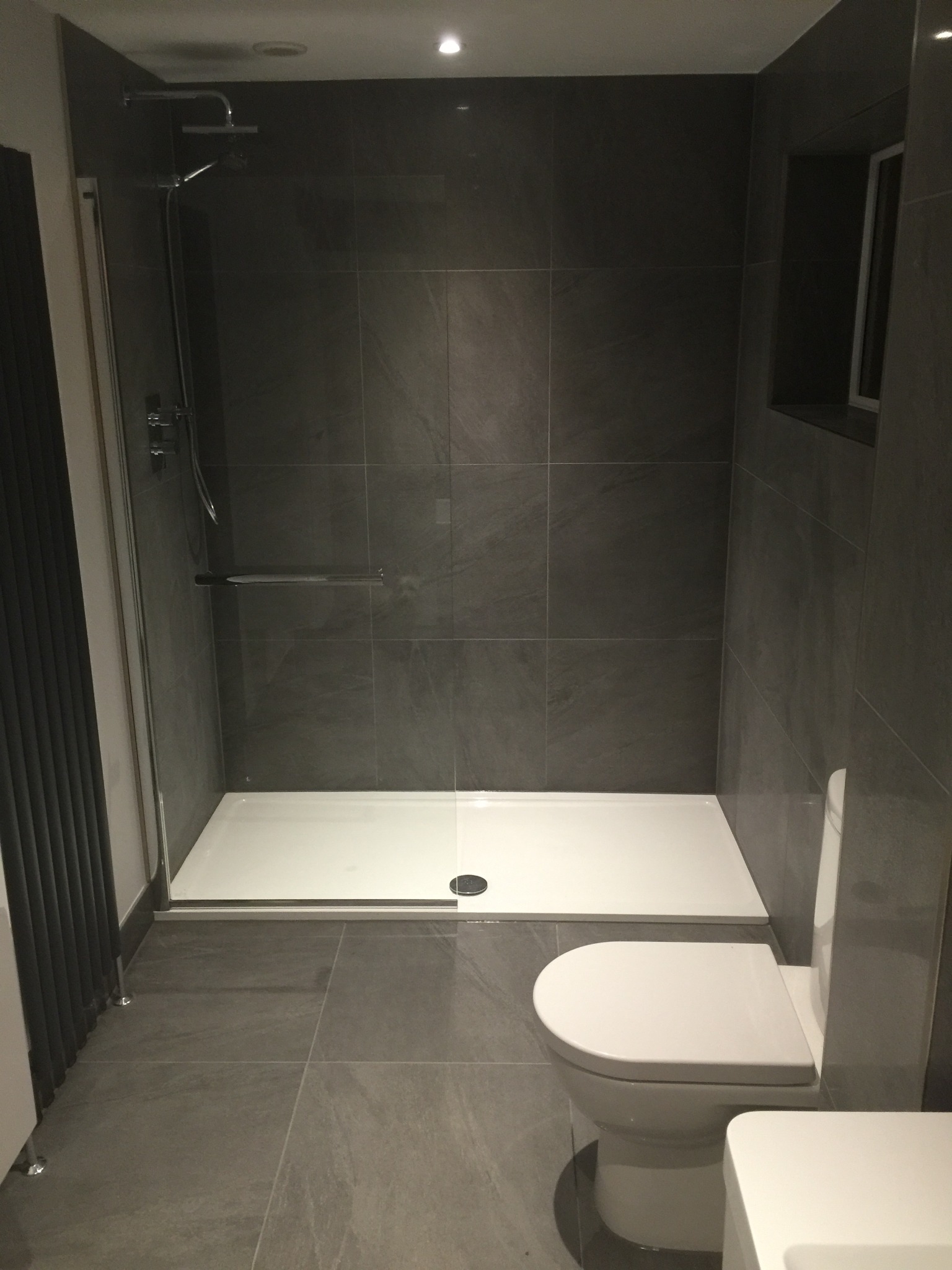 Nowoczesna łazienka w odcieniach szarości z kabiną prysznicową typu walk-in, umywalką i toaletą. Ściany i podłoga wyłożone dużymi, kwadratowymi płytkami imitującymi kamień. W kabinie prysznicowej...