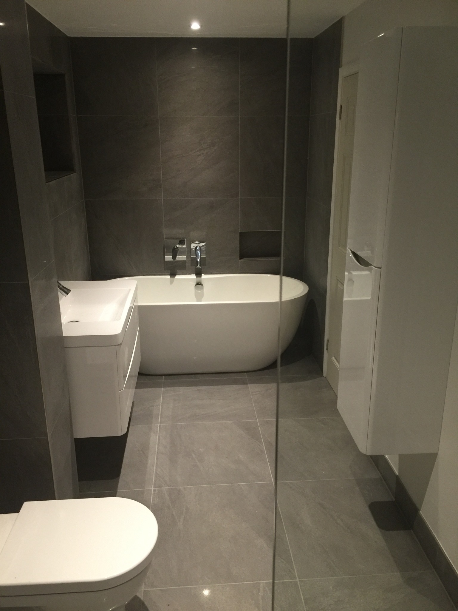 Nowoczesna łazienka z szarymi płytkami, białą wanną wolnostojącą, umywalką z szafką i podwieszaną szafką, oświetlona punktowymi lampami sufitowymi, oddzielona szklaną ścianką.