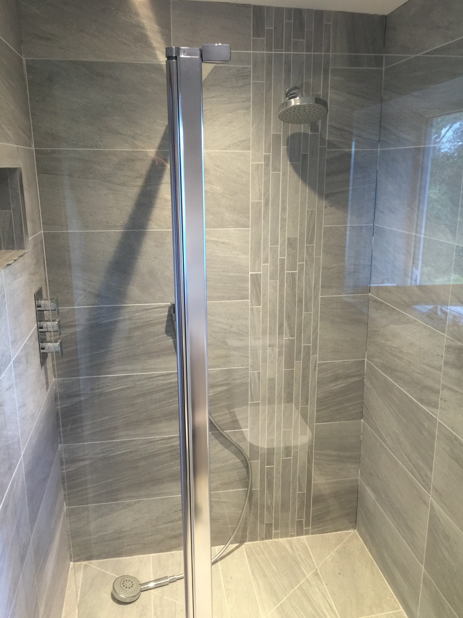 Szara łazienka z kabiną prysznicową ze srebrnymi ramami i panelami sterującymi, wykończona płytkami imitującymi kamień z akcentem pionowych płytek.