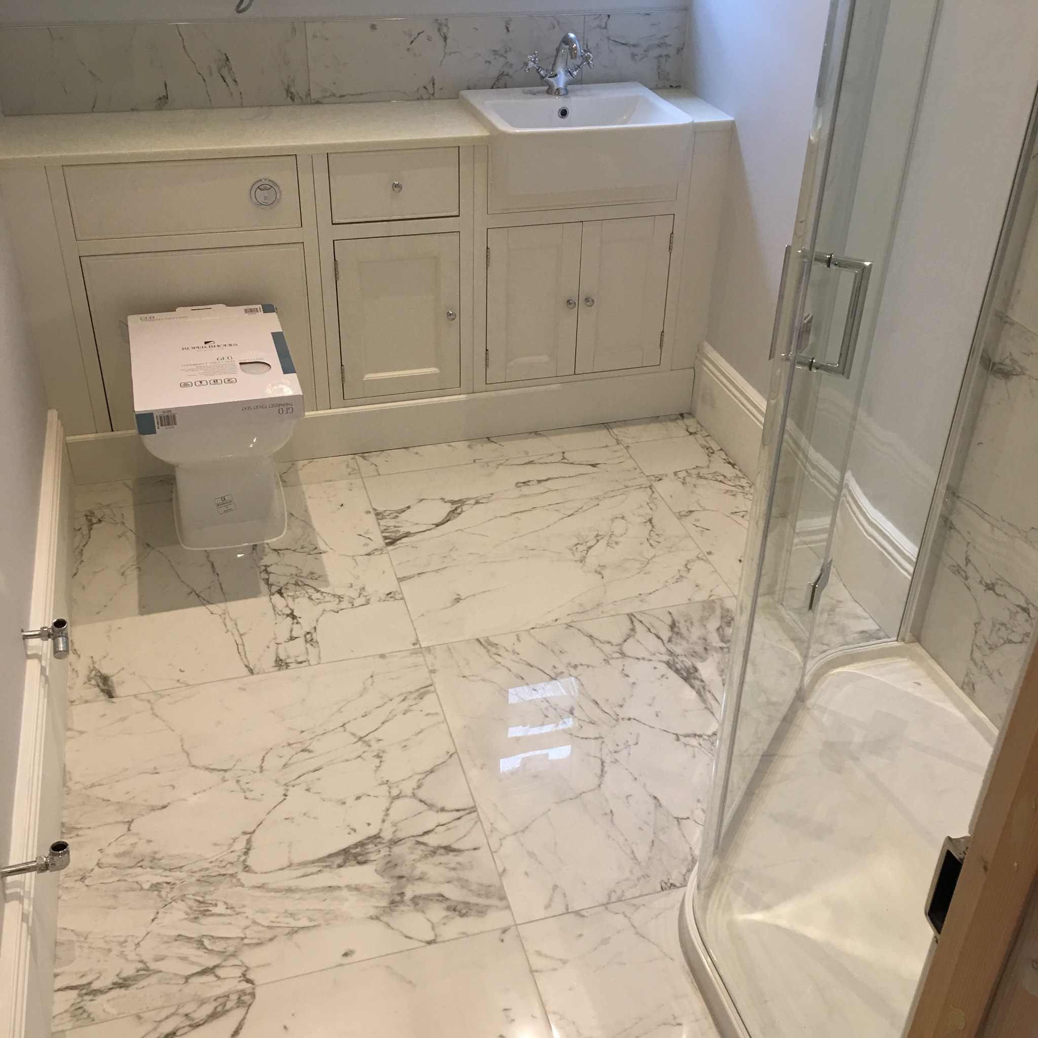 Wykończona łazienka w domu prywatnym z białymi marmurowymi płytkami podłogowymi, zabudowaną szafką pod umywalką, kabiną prysznicową i toaletą w kartonie marki Roper Rhodes.
