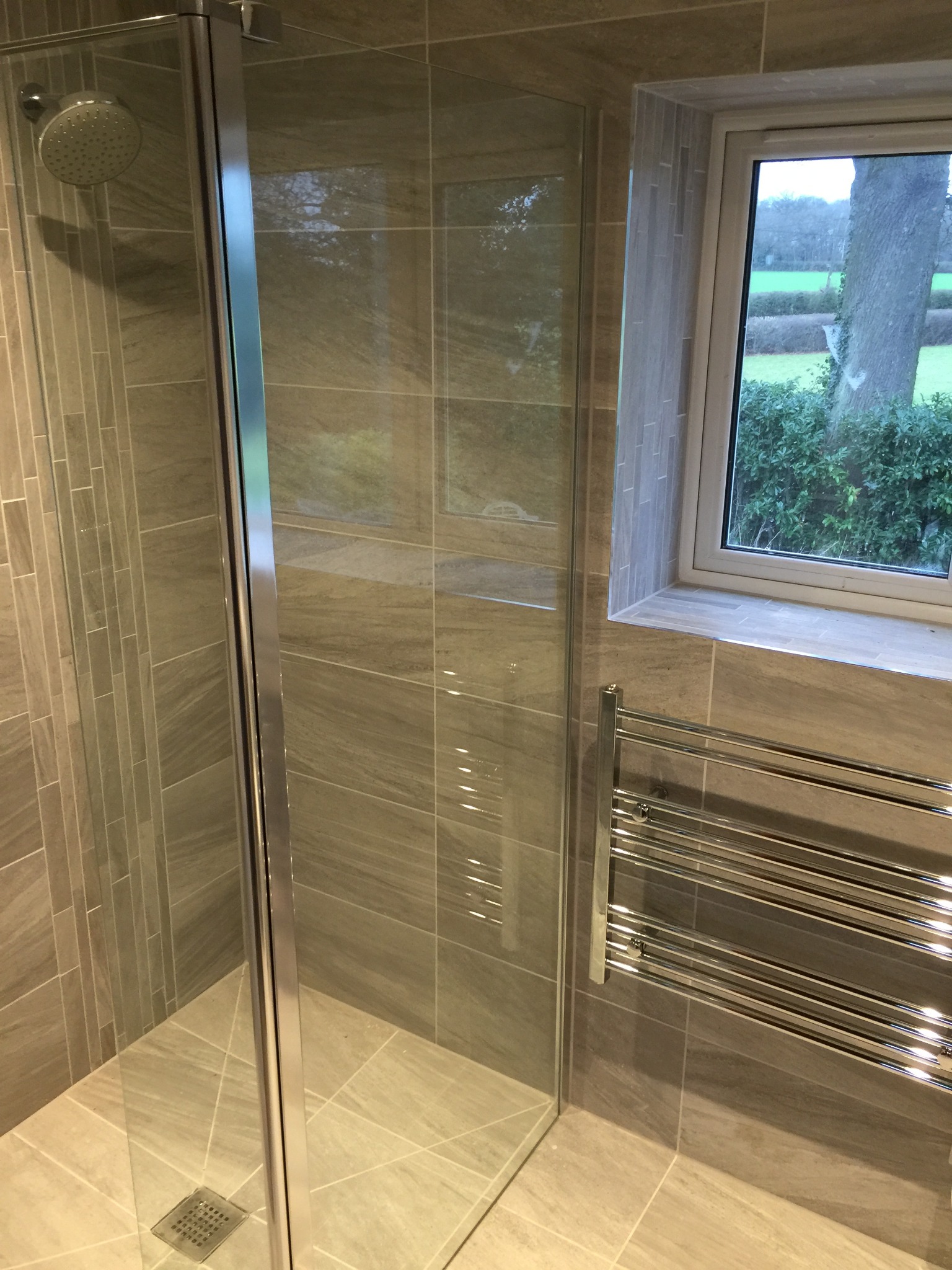 Nowoczesna kabina prysznicowa z przeszklonymi drzwiami i odpływem liniowym, obok okno z widokiem na zieleń, grzejnik drabinkowy na ścianie wyłożonej płytkami w odcieniach szarości.