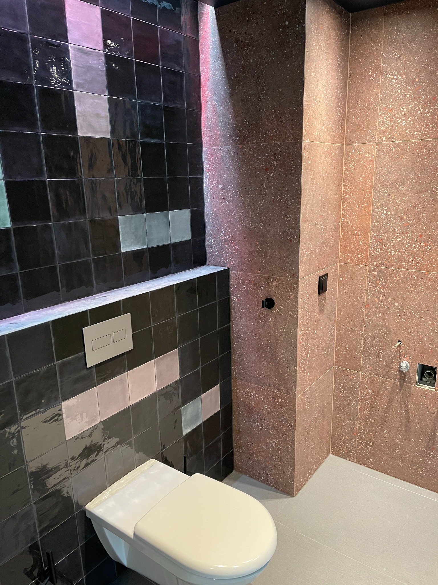 Nowoczesna łazienka w trakcie wykańczania, z czarnymi błyszczącymi płytkami na ścianie, fragmentem toalety i ścianą wyłożoną płytkami w odcieniach czerwieni z drobnymi wzorami.