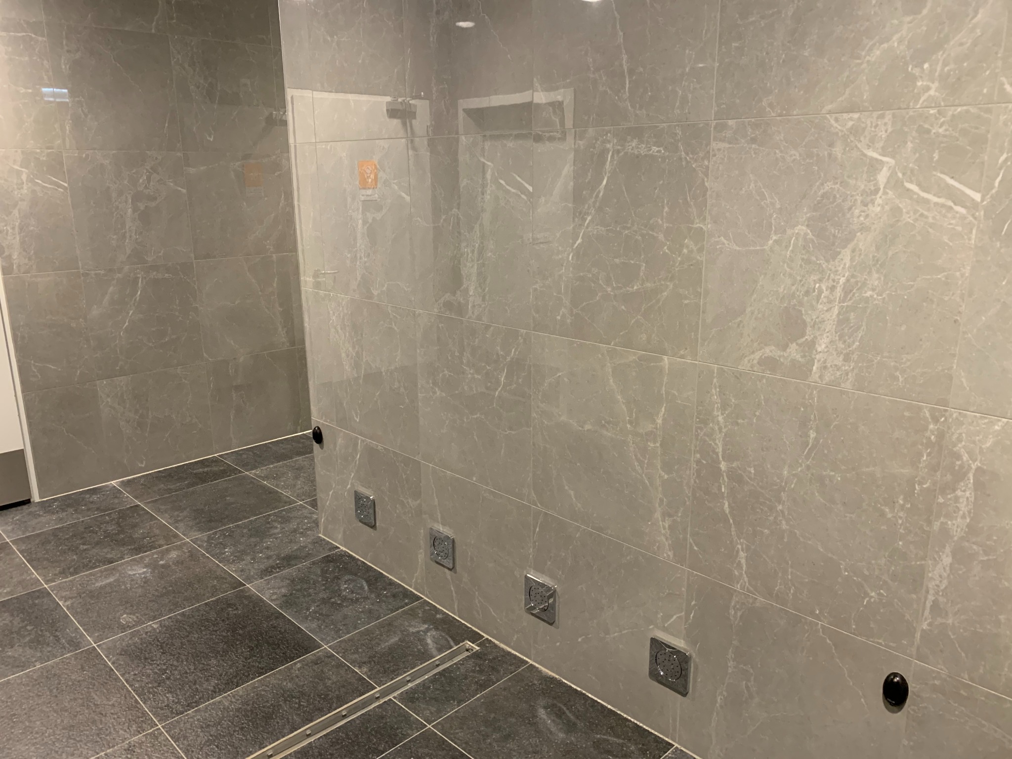 Wykończona łazienka z szarymi, marmurkowymi płytkami na ścianach i ciemnoszarą podłogą. Widoczne metalowe odpływy liniowe i punkty podłączenia wody.