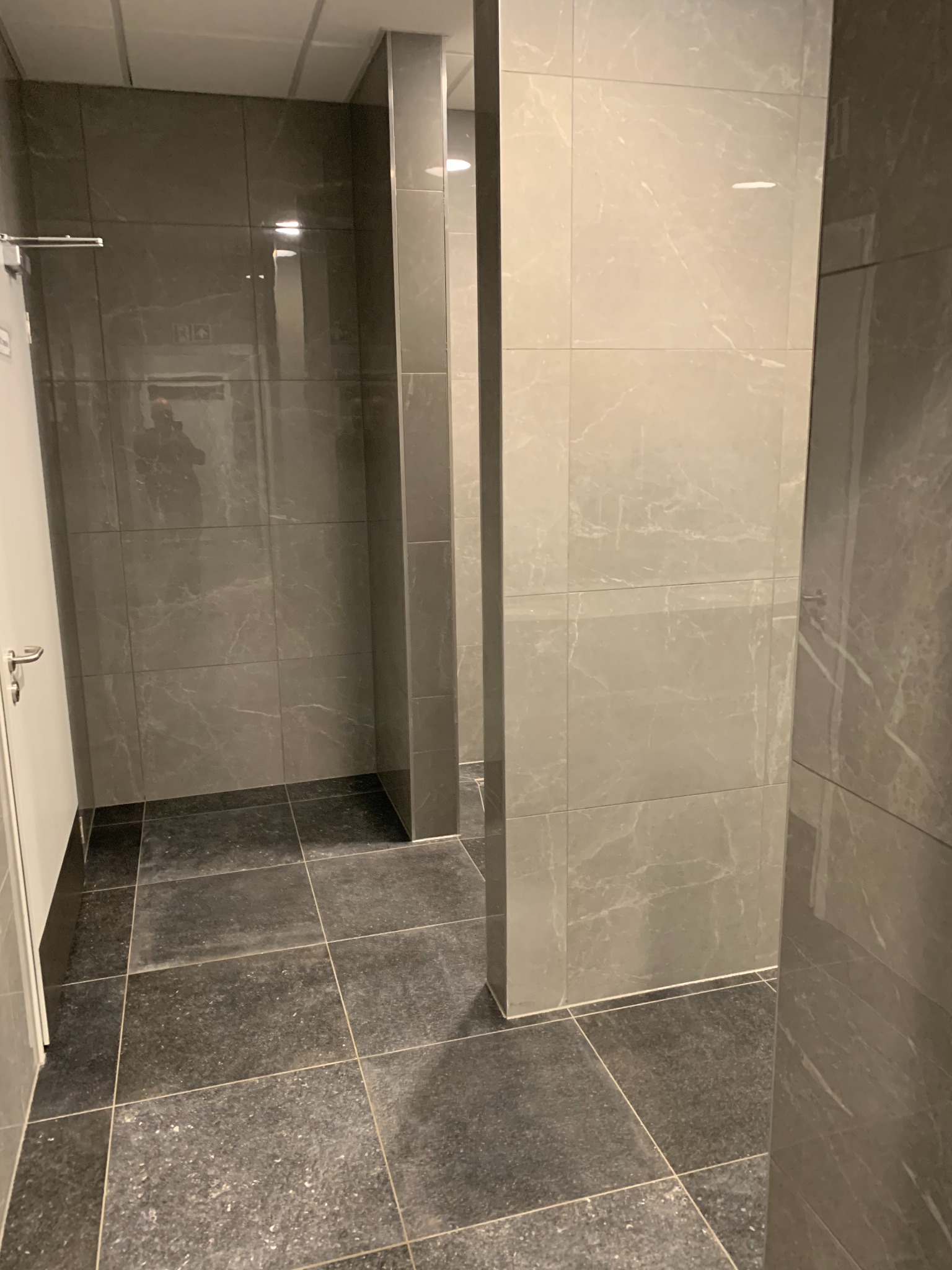 Wykończona łazienka w szarych tonacjach z płytkami imitującymi marmur na ścianach i ciemnymi płytkami podłogowymi ułożonymi w kwadraty, widoczna kabina prysznicowa z szklanymi drzwiami.