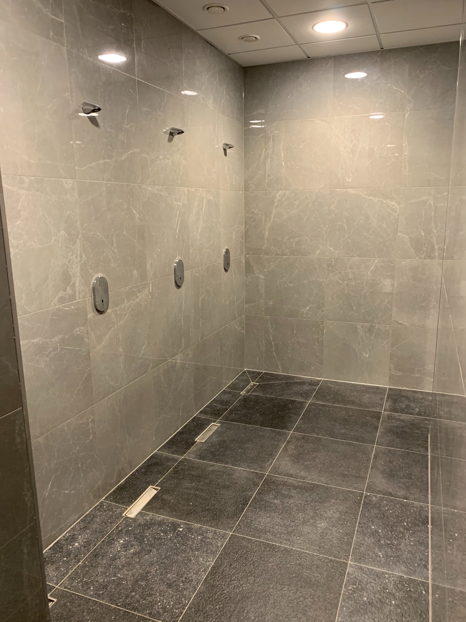 Wykończona kabina prysznicowa z szarymi płytkami imitującymi marmur na ścianach i ciemnoszarą podłogą z odpływami liniowymi. Na ścianie widoczne chromowane wieszaki.