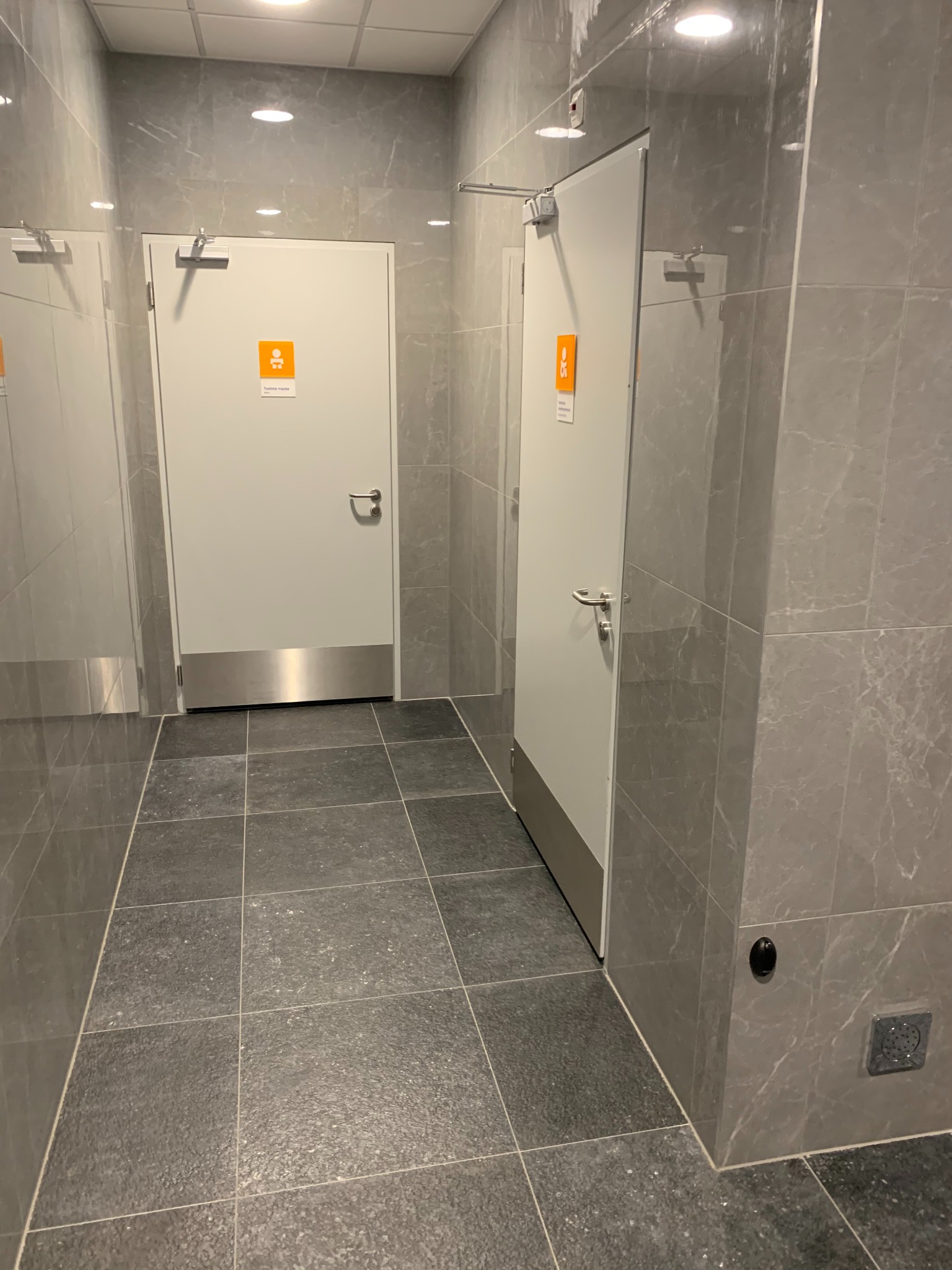 Wykończona łazienka publiczna z szarymi płytkami imitującymi marmur na ścianach i podłodze, białe drzwi z oznaczeniami toalet.