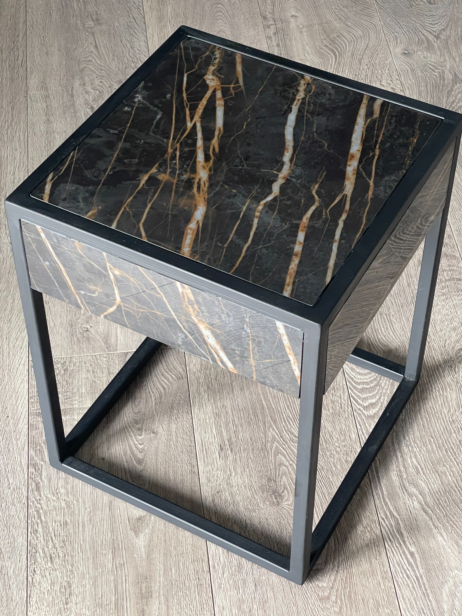 Stolik nocny z blatem imitującym czarny marmur z wyraźnymi złotymi żyłami, na metalowej ramie w kolorze grafitowym, ustawiony na podłodze z paneli imitujących drewno.