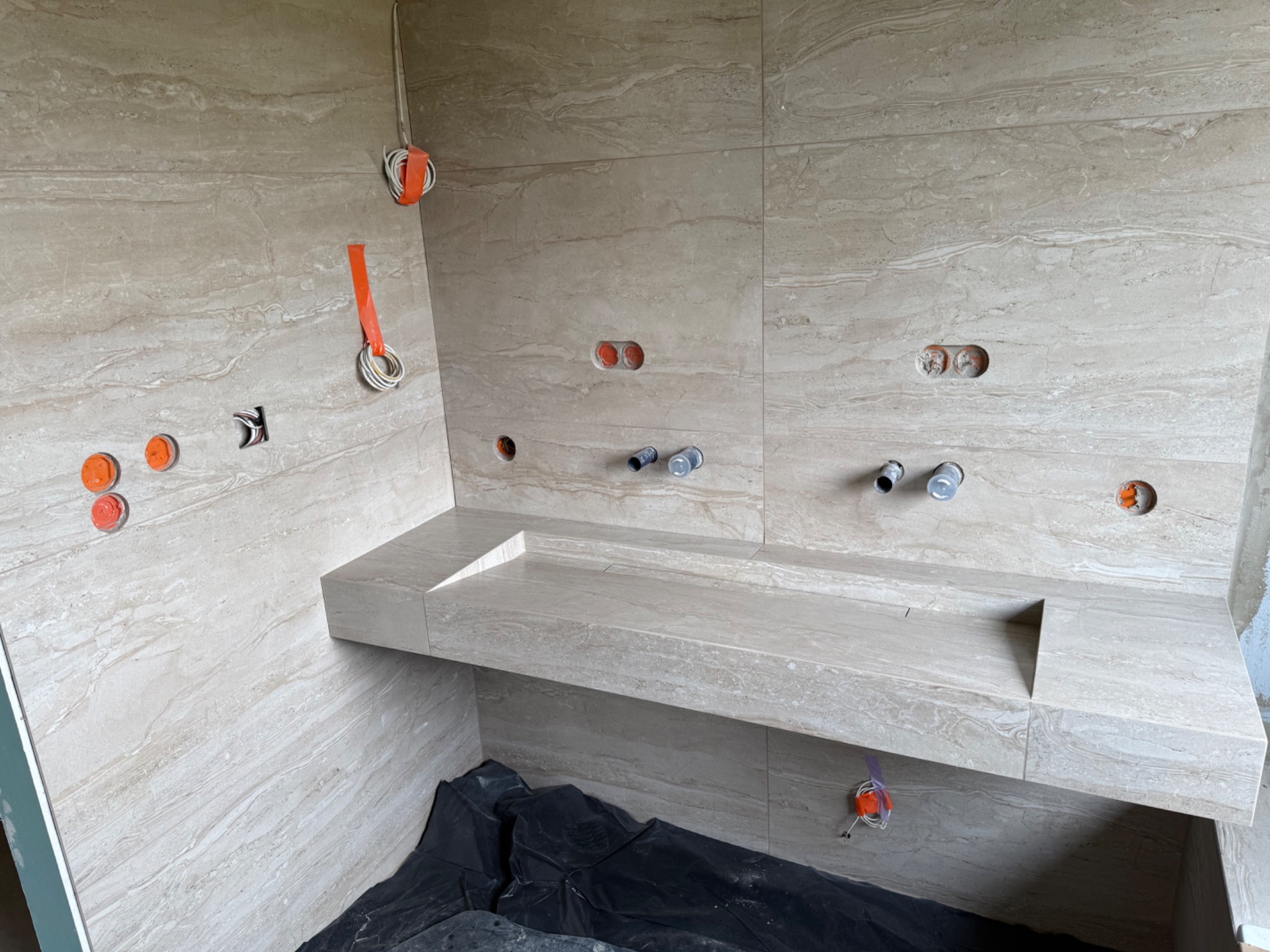 Beżowa łazienka w trakcie wykańczania: marmurowe płytki, zabudowana umywalka, widoczne instalacje elektryczne i hydrauliczne. Wnętrze w stylu minimalistycznym.