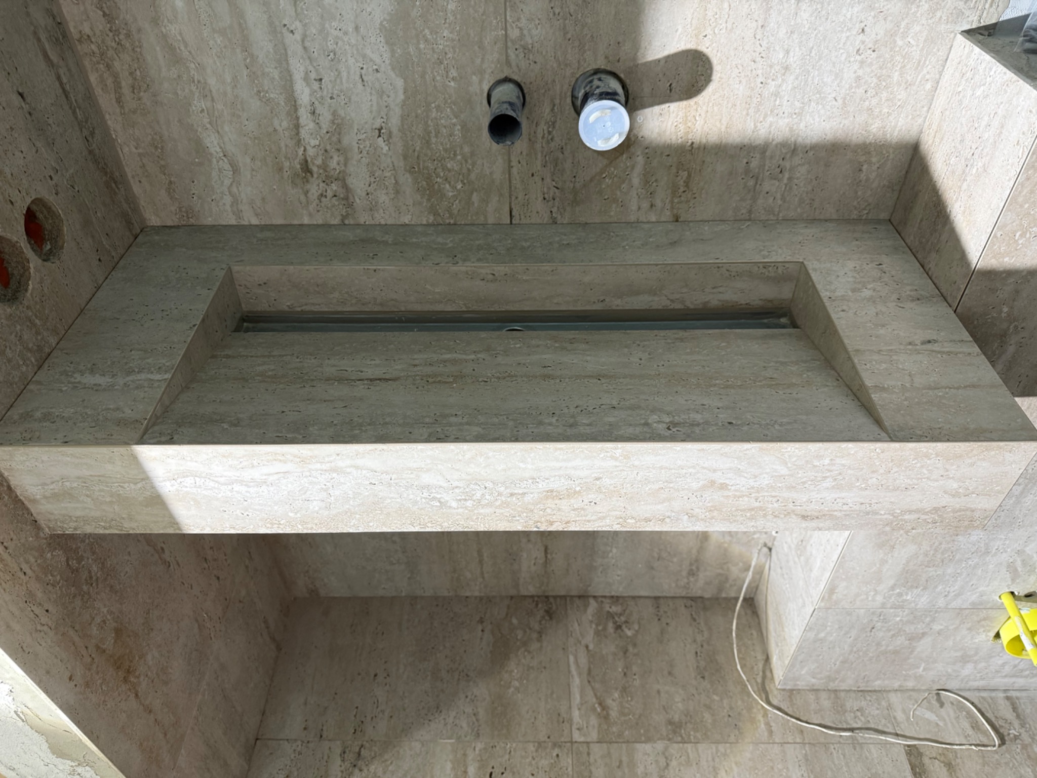 Wykończona umywalka z trawertynu z odpływem liniowym, widoczne rury doprowadzające wodę i otwory na instalację elektryczną. Minimalistyczny design łazienki.