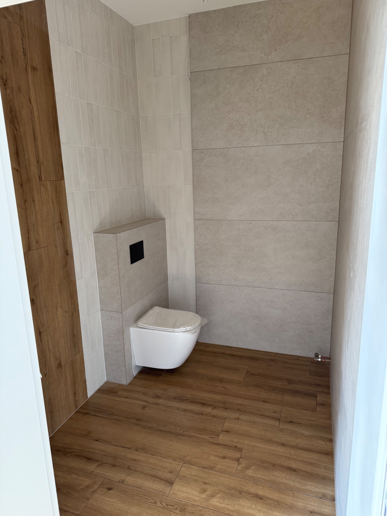 Nowoczesna łazienka z drewnopodobną podłogą, beżowymi płytkami na ścianach i podwieszaną toaletą. Minimalistyczny design, ciepłe odcienie drewna i kamienia.