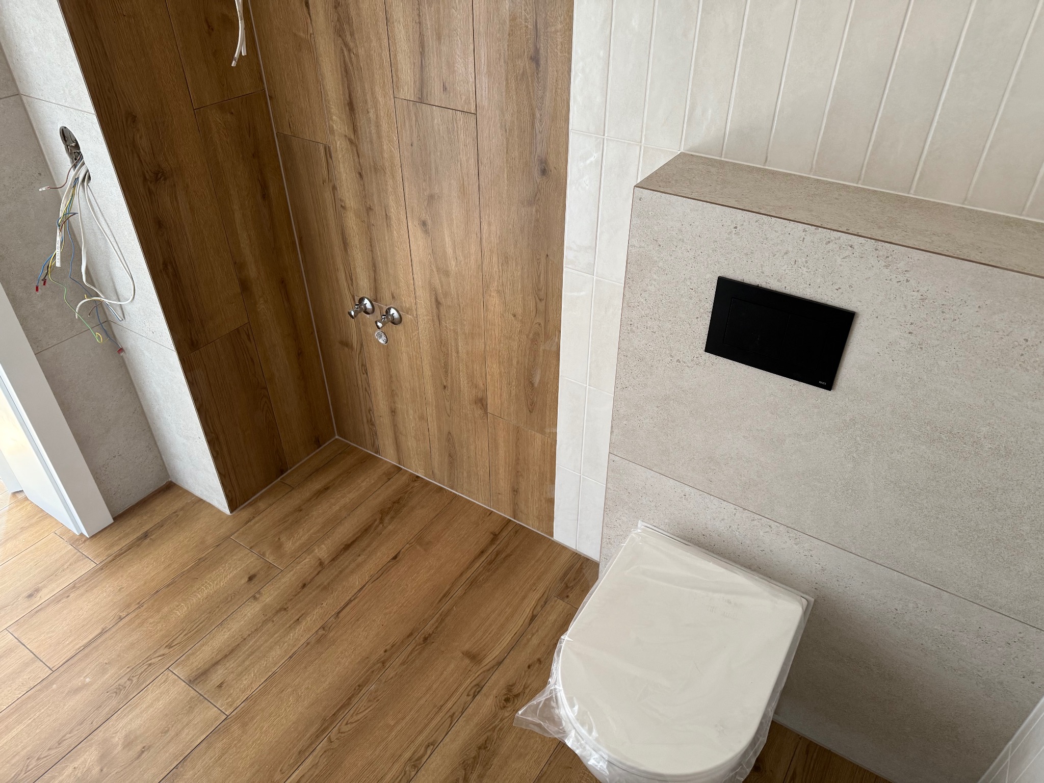 Nowoczesna łazienka z drewnopodobnymi płytkami na podłodze i ścianie prysznicowej, oraz beżowymi płytkami na pozostałych ścianach. Wisząca toaleta w folii.