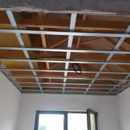 Montaż metalowej konstrukcji sufitu podwieszanego z widoczną izolacją i drewnianą konstrukcją nośną powyżej, w surowym stanie wykończenia wnętrza z oknem.