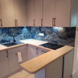 Nowoczesna kuchnia w odcieniach szarości z blatem z jasnego drewna i dekoracyjnym panelem ściennym w odcieniach błękitu i szarości imitującym marmur. Czarny zlew i płyta indukcyjna wkomponowane...