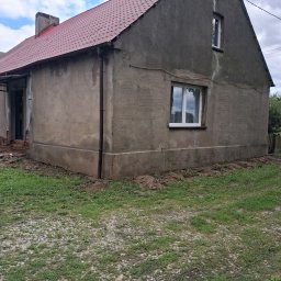 USŁUGI OGÓLNOBUDOWLANE JAROSŁAW STRAUCHMANN - Szary dom jednorodzinny z czerwoną dachówką, częściowo otynkowany. Widoczne ślady prac remontowych wokół budynku i świeżo wstawione okna.