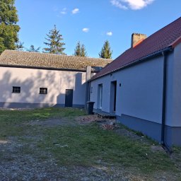 USŁUGI OGÓLNOBUDOWLANE JAROSŁAW STRAUCHMANN - Odnowiony budynek gospodarczy z nową elewacją w odcieniach szarości, z widocznym fragmentem dachu z czerwonej dachówki i zielonym otoczeniem. Widok z boku, w tle drzewa.