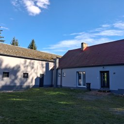 USŁUGI OGÓLNOBUDOWLANE JAROSŁAW STRAUCHMANN - Odnowiony budynek gospodarczy z nową elewacją w odcieniach szarości i czerwoną dachówką, widoczny fragment niebieskiego samochodu i trawnik w słoneczny dzień.