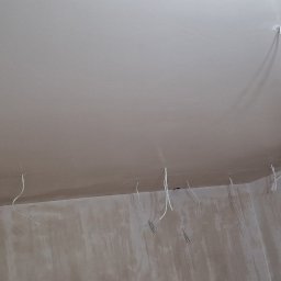 USŁUGI OGÓLNOBUDOWLANE JAROSŁAW STRAUCHMANN - Wnętrze w trakcie prac wykończeniowych: gładki sufit po szpachlowaniu, wystające przewody elektryczne, ściany przygotowane do malowania. Widok z dołu, ukośne ujęcie.
