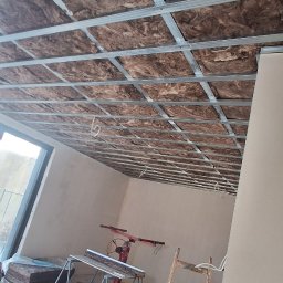 USŁUGI OGÓLNOBUDOWLANE JAROSŁAW STRAUCHMANN - Poddasze w trakcie ocieplania: wełna mineralna między metalowymi profilami, widoczne narzędzia i drabina. Prace wykończeniowe w surowym wnętrzu z oknem.