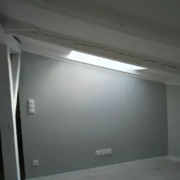 Poddasze po malowaniu na szaro, widoczne białe belki stropowe, okno dachowe i gniazdka elektryczne przy podłodze.