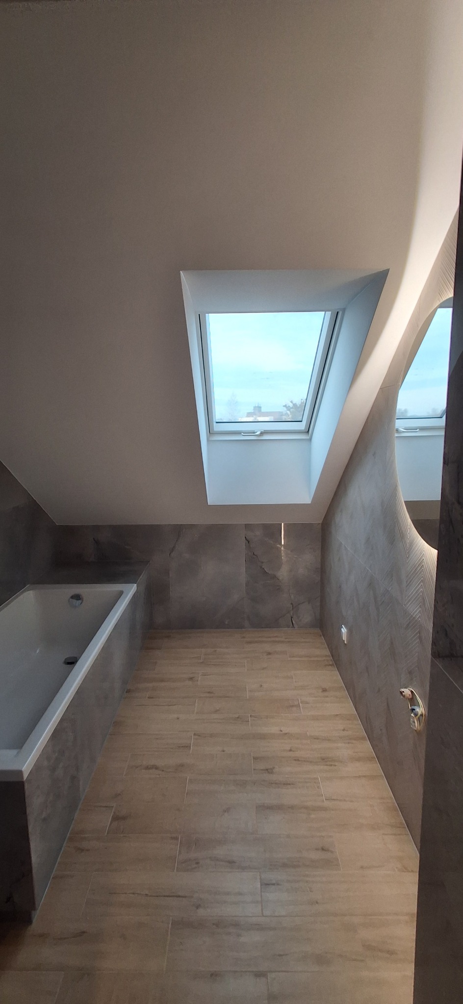 Nowoczesna łazienka na poddaszu z oknem dachowym, wykończona płytkami imitującymi drewno i kamień. Minimalistyczny design, jasne wnętrze.