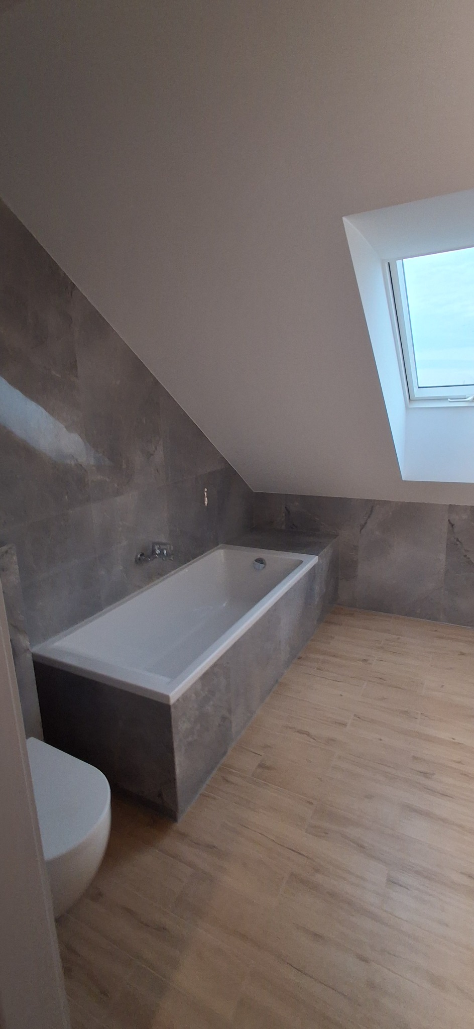 Nowoczesna łazienka na poddaszu z wanną obudowaną szarą płytką imitującą marmur, drewnopodobną podłogą i oknem dachowym. Minimalistyczny design.