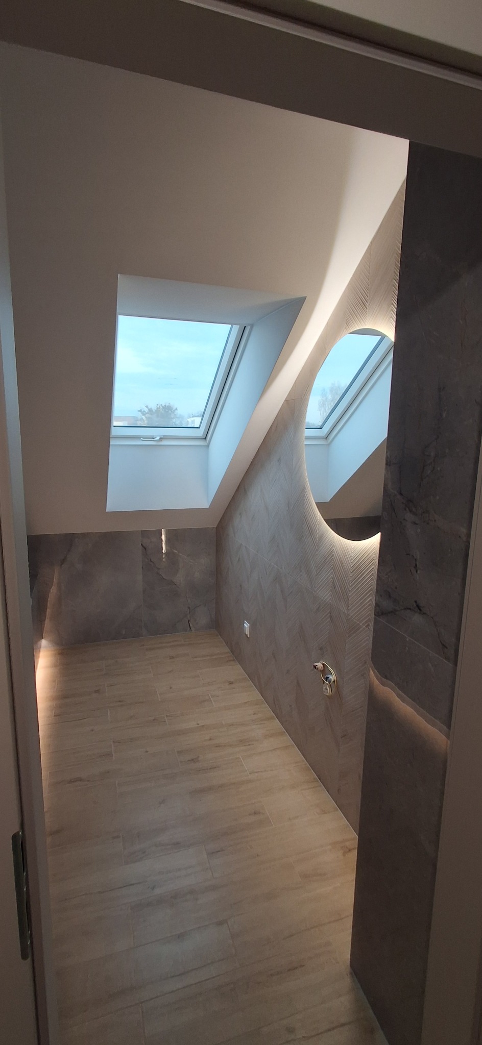 Nowoczesna łazienka na poddaszu z oknem dachowym, okrągłym lustrem z podświetleniem LED i płytkami imitującymi drewno na podłodze i ścianach.