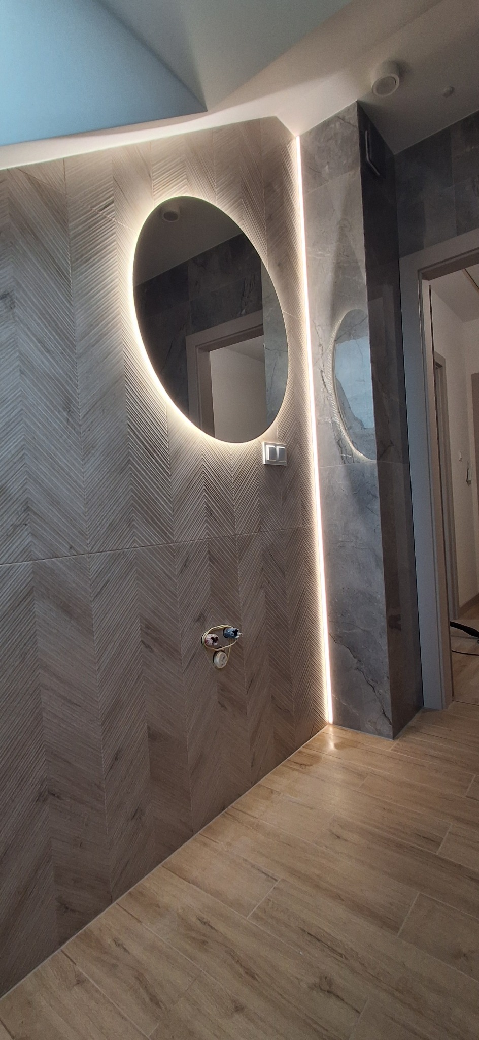 Nowoczesna łazienka z podświetlanym lustrem i płytkami w jodełkę. Wykończenie w odcieniach szarości i beżu, drewnopodobna podłoga. Minimalistyczny design.