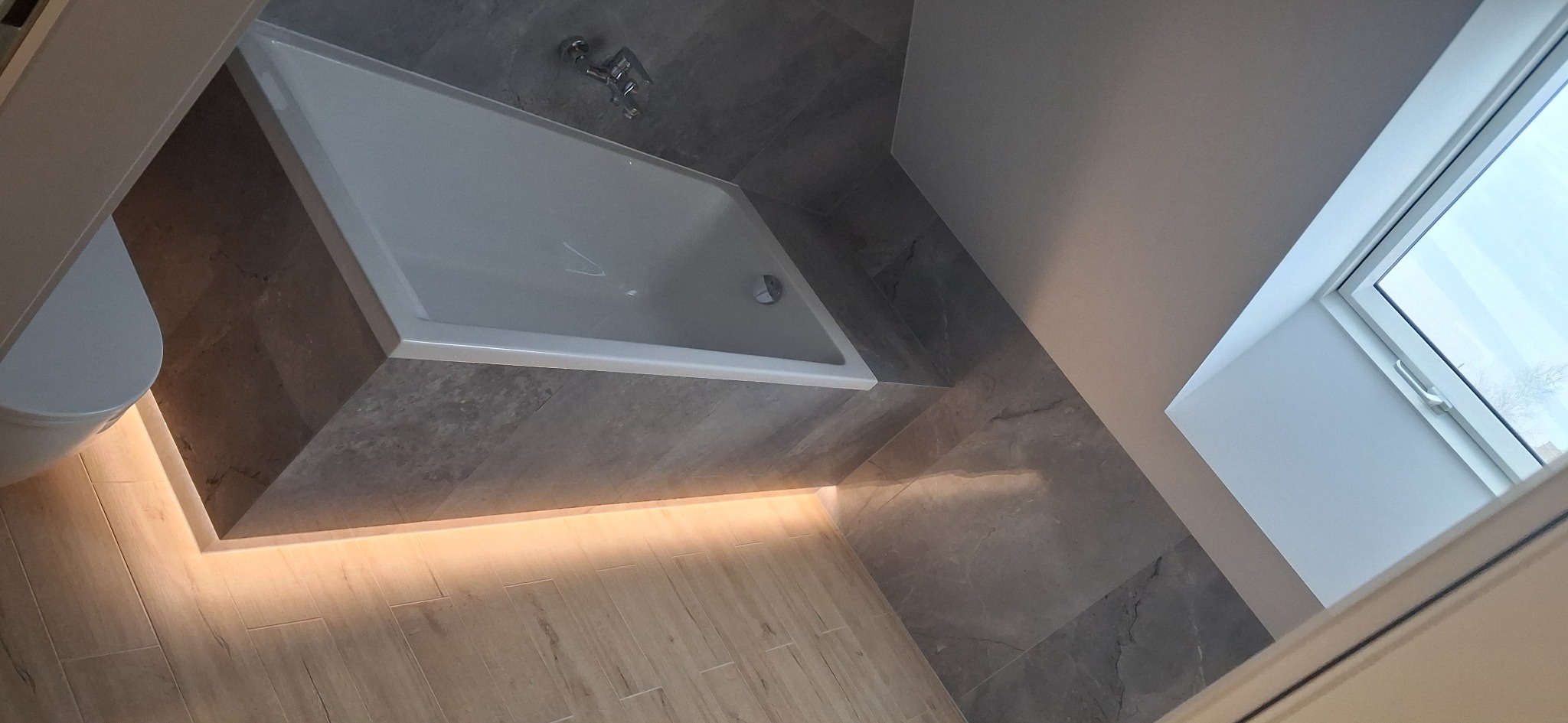 Nowoczesna łazienka z podświetlaną wanną, szarą glazurą i oknem dachowym. Ujęcie z góry podkreśla minimalistyczny design i dbałość o detale.