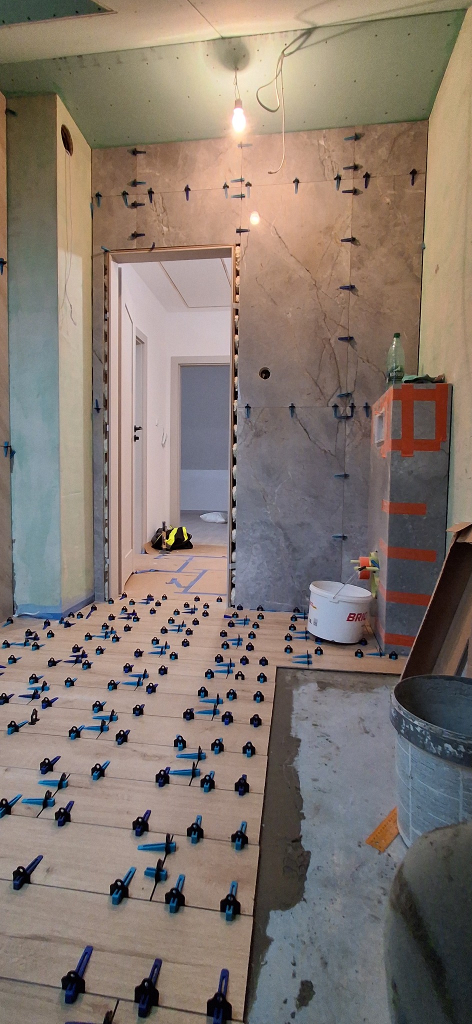 Układanie płytek podłogowych i ściennych w łazience z użyciem systemu poziomowania. Widoczne płytki imitujące drewno i marmur, narzędzia glazurnicze i wiadro z klejem.