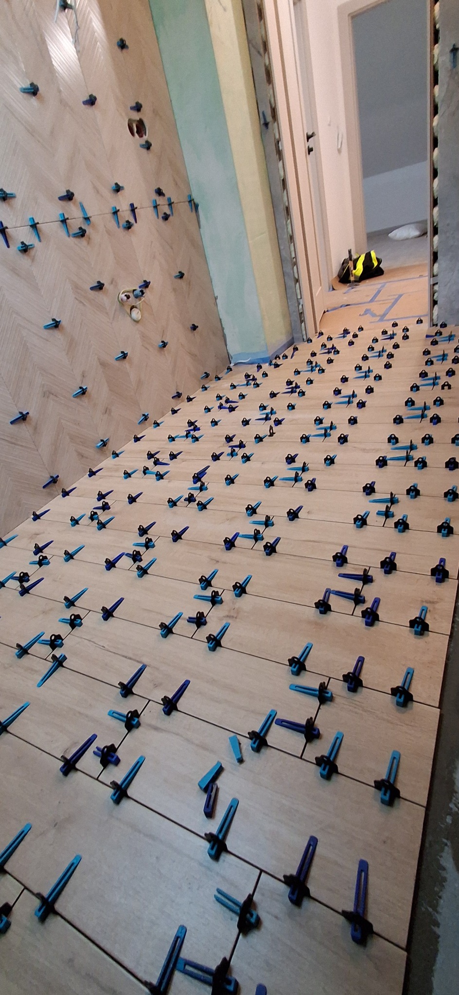 Ułożone płytki podłogowe w jodełkę, z niebieskimi krzyżykami dystansowymi, widok z góry, w tle otwarte drzwi i torba robocza.