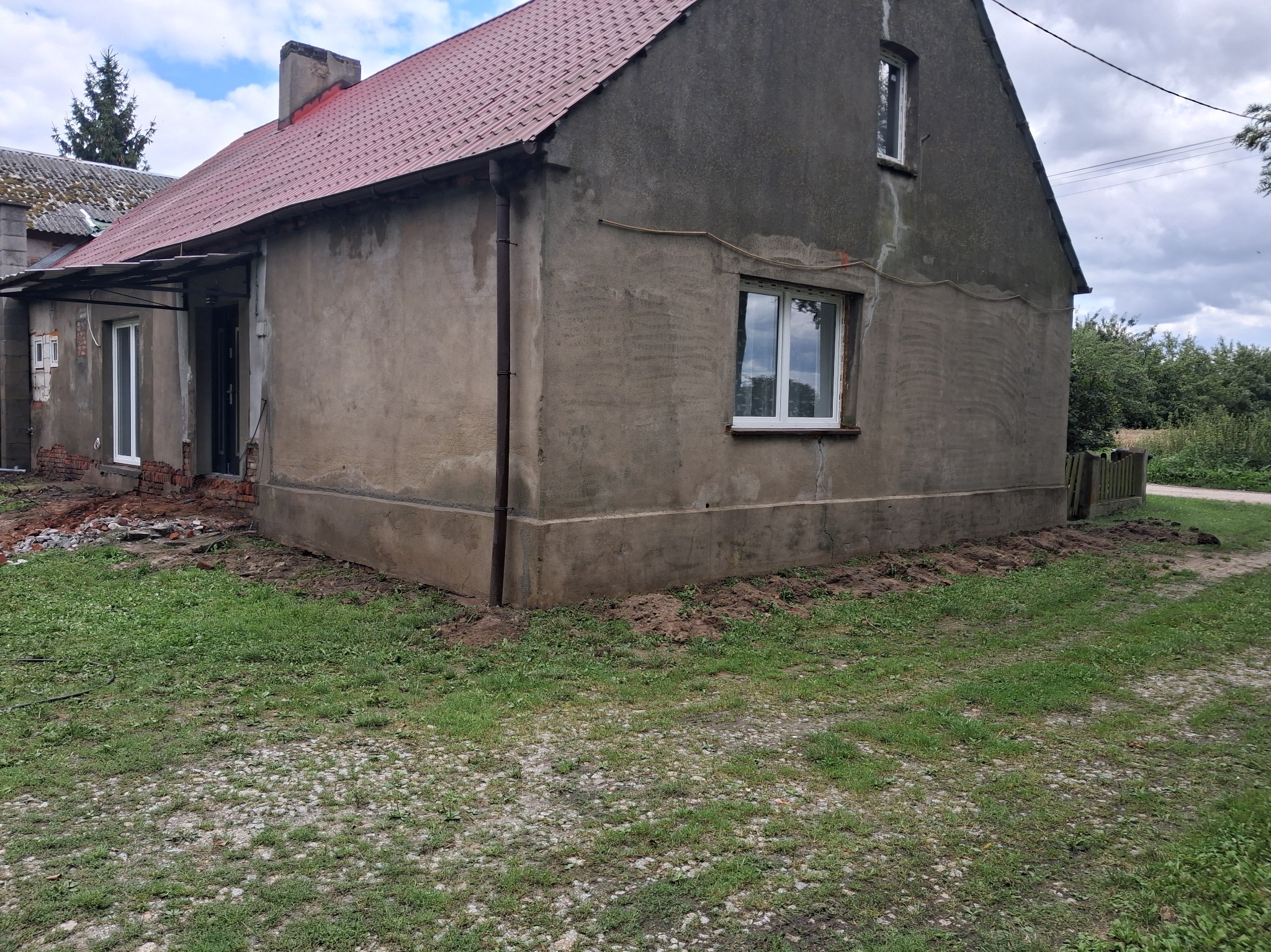Szary dom jednorodzinny z czerwoną dachówką, częściowo otynkowany. Widoczne ślady prac remontowych wokół budynku i świeżo wstawione okna.