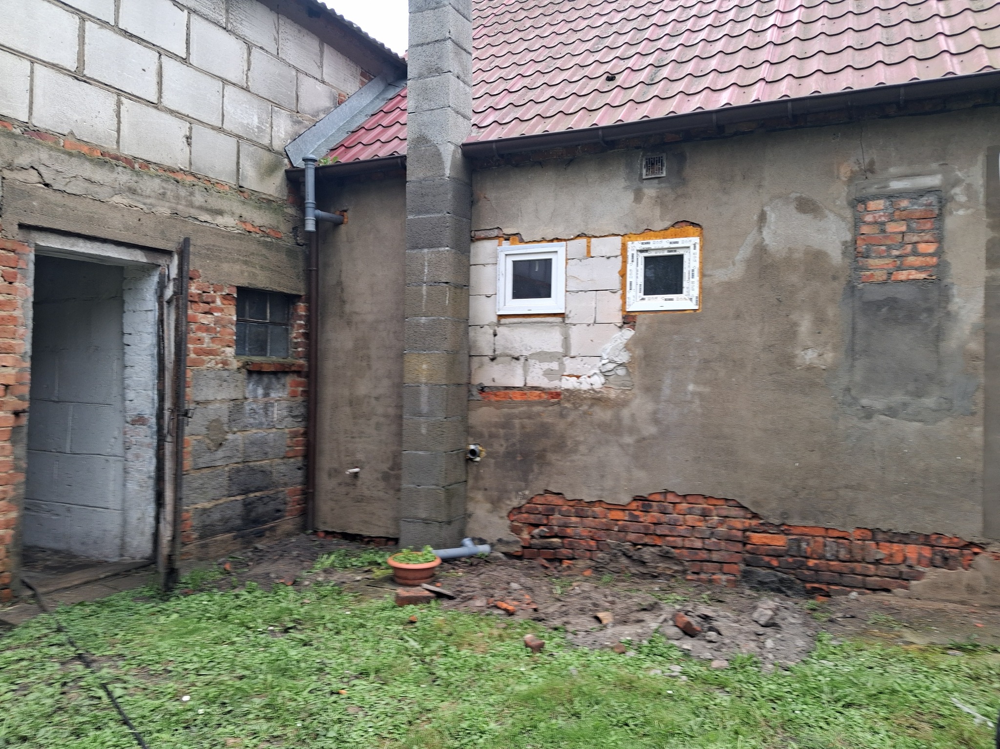 Fragment budynku z widocznymi śladami napraw i wymiany okien. Częściowo odnowiona elewacja z ceglanymi elementami i nowymi oknami w białych ramach.