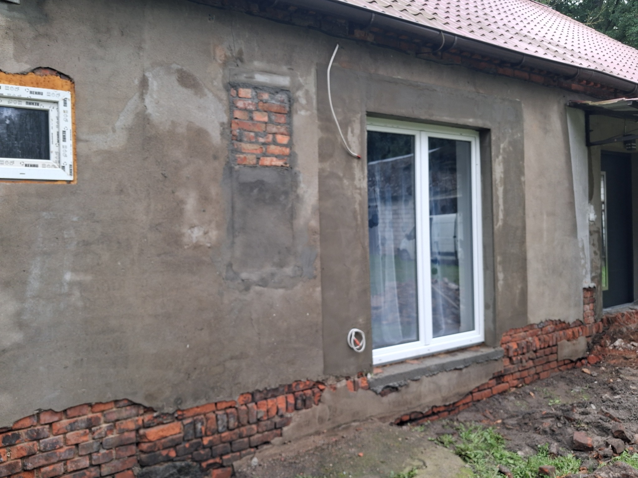 Fragment elewacji domu w trakcie prac remontowych: widoczne nowe okna, fragmenty odsłoniętej cegły i niedokończone tynki. Prace murarskie i tynkarskie w toku.