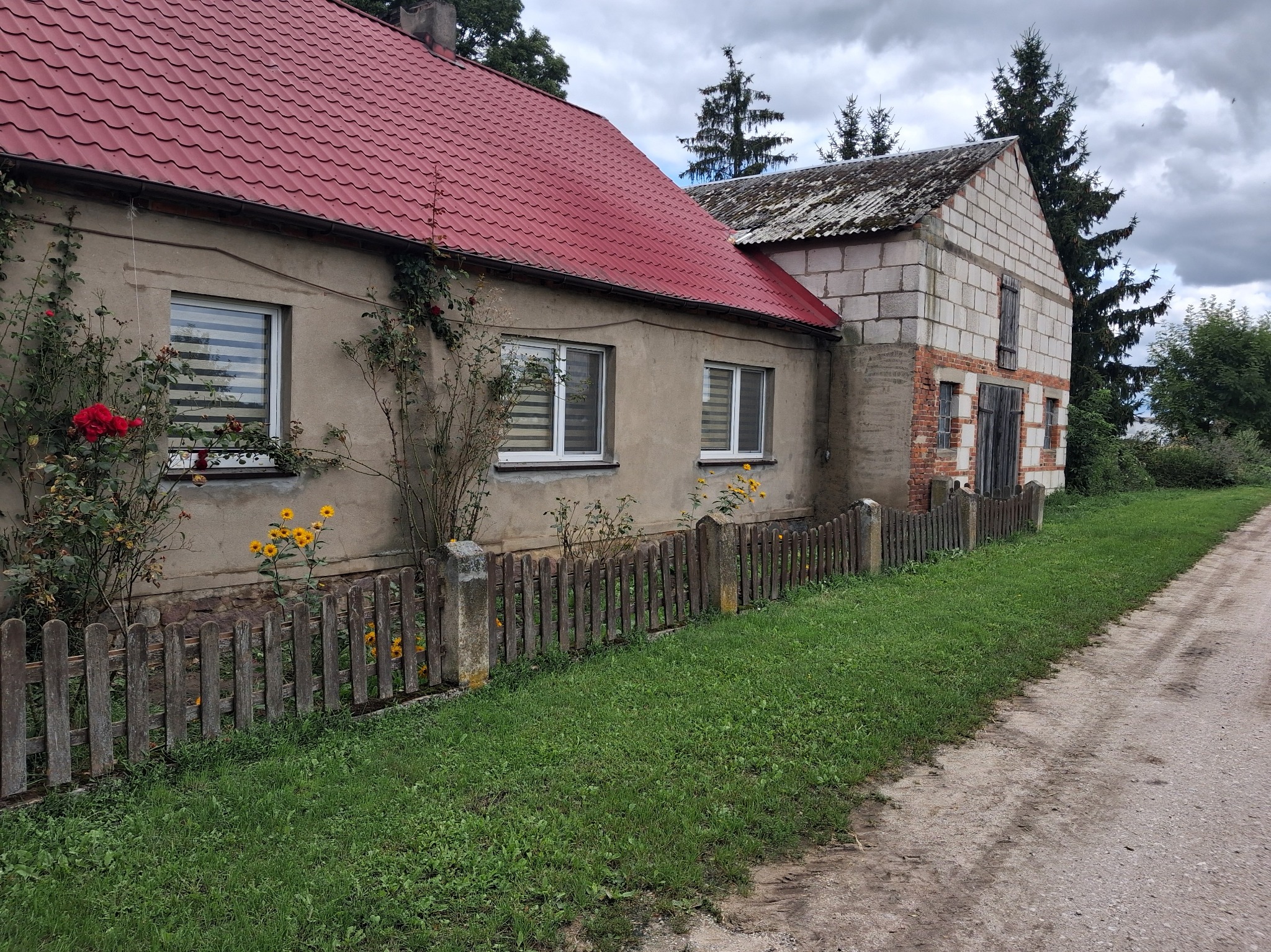 Dom z czerwonym dachem, drewnianym płotem i kwitnącymi kwiatami w ogrodzie. Widoczne okna z żaluzjami i fragment dobudówki z białego pustaka.