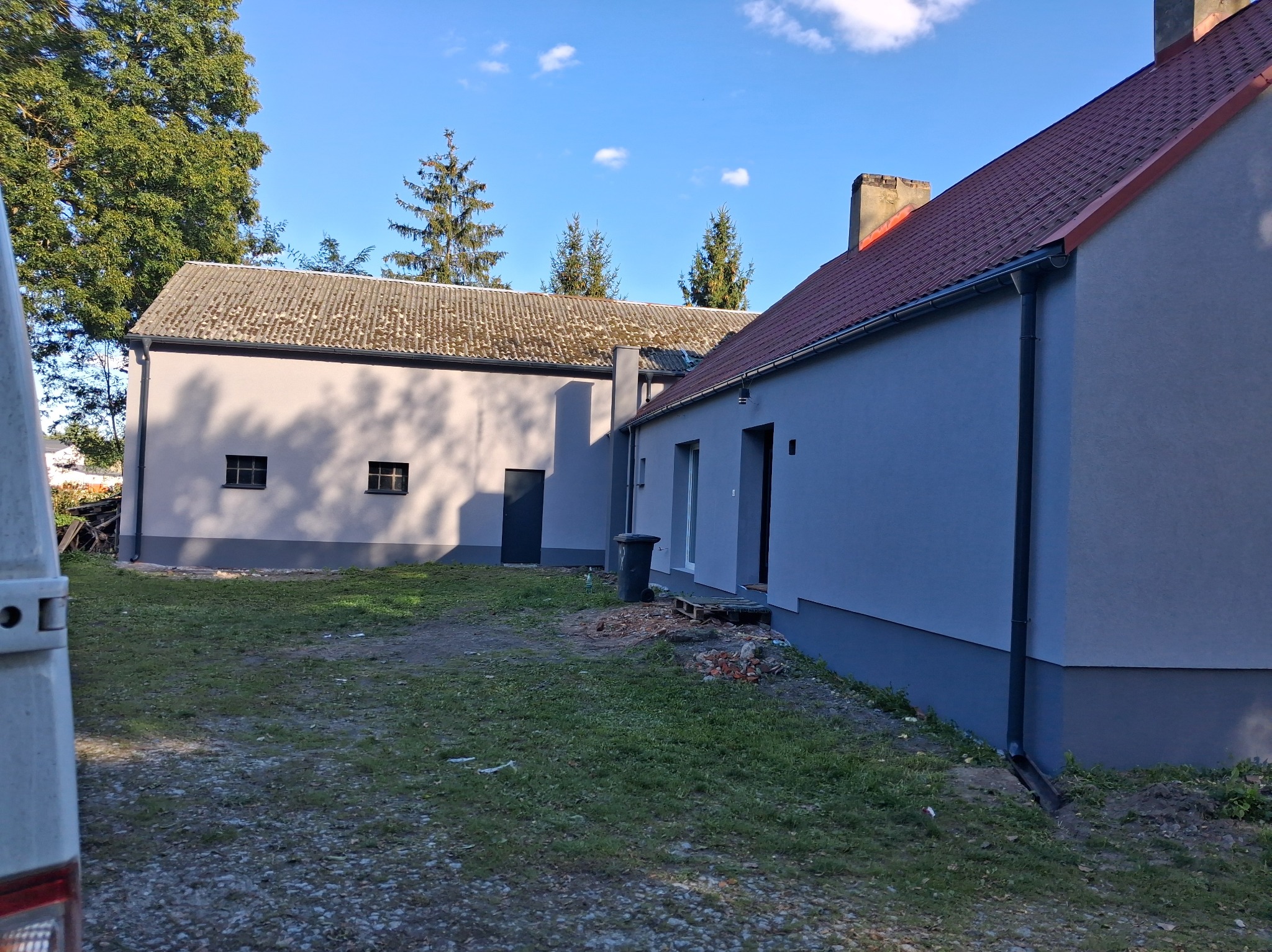 Odnowiony budynek gospodarczy z nową elewacją w odcieniach szarości, z widocznym fragmentem dachu z czerwonej dachówki i zielonym otoczeniem. Widok z boku, w tle drzewa.