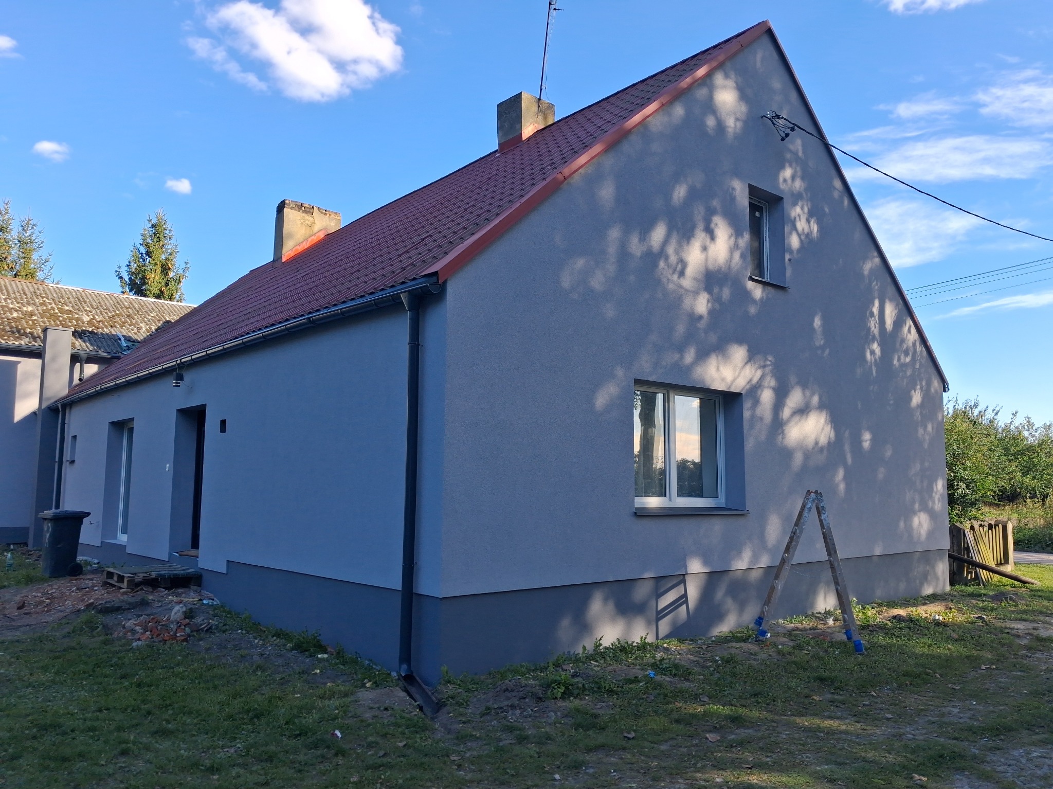 Odnowiony dom jednorodzinny z szarą elewacją i czerwoną dachówką w słoneczny dzień. Widoczne drzewa rzucające cień na ścianę i drabina oparta o budynek.