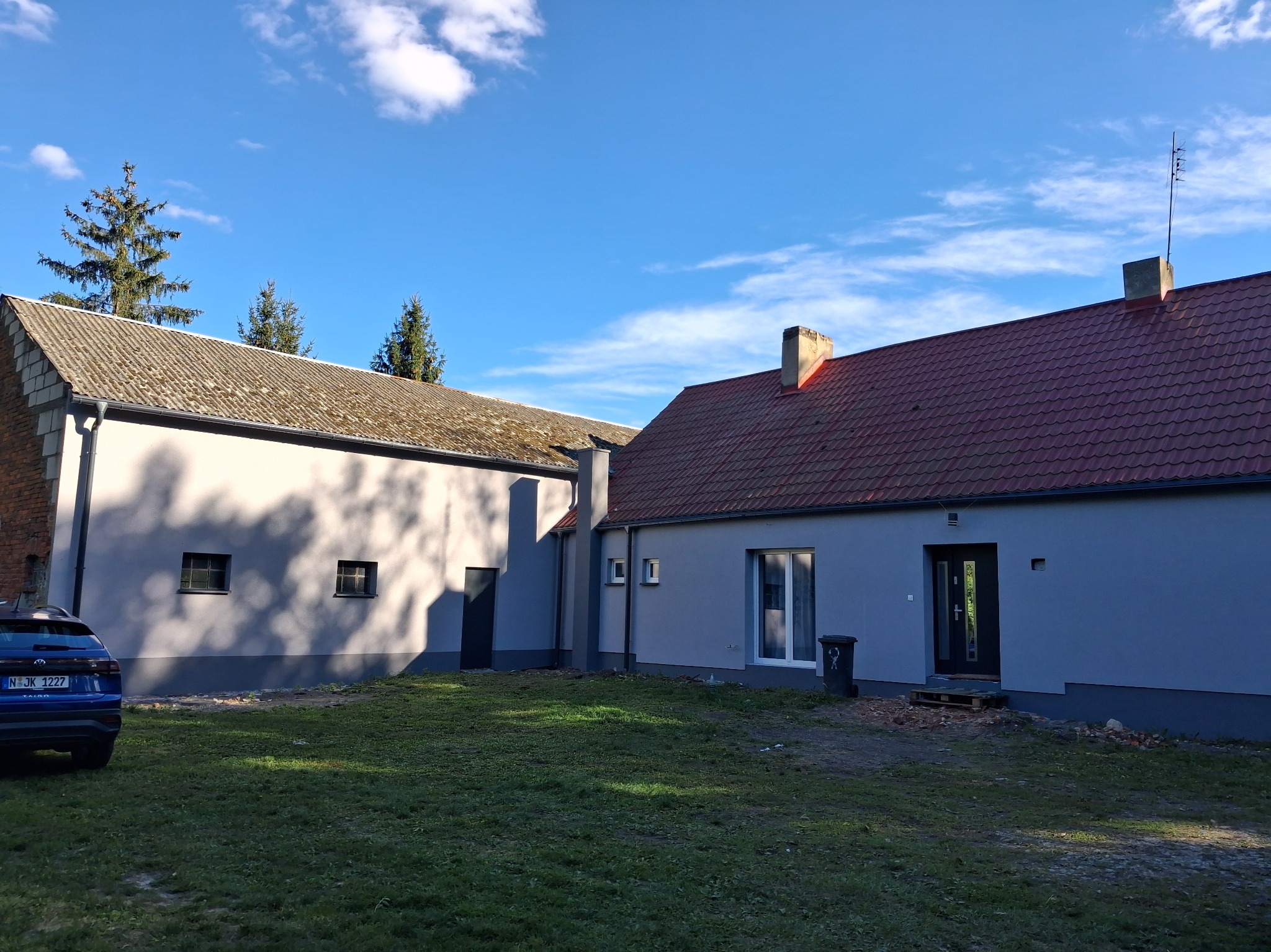 Odnowiony budynek gospodarczy z nową elewacją w odcieniach szarości i czerwoną dachówką, widoczny fragment niebieskiego samochodu i trawnik w słoneczny dzień.