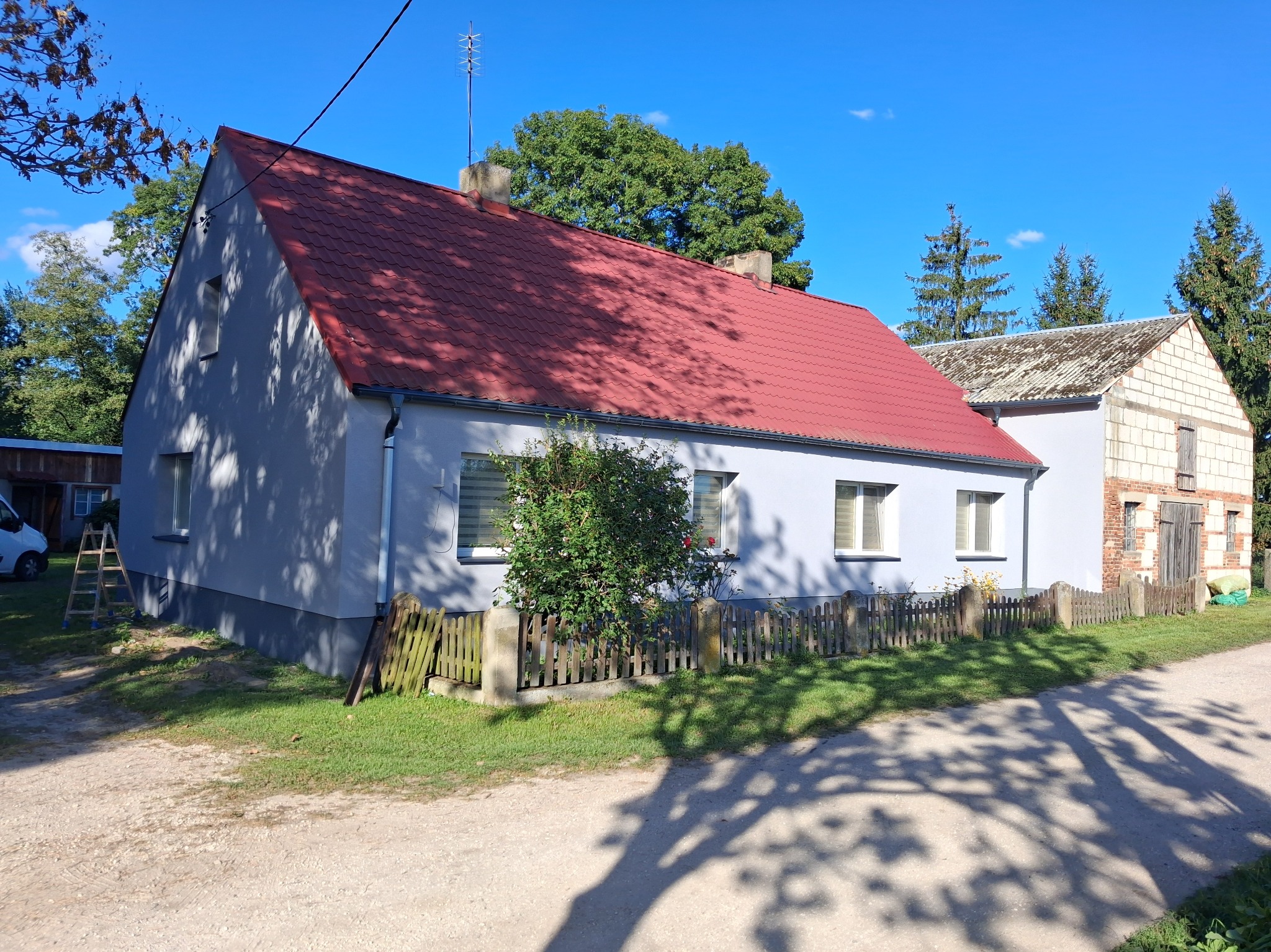 Odnowiony dom jednorodzinny z czerwoną dachówką i jasną elewacją, otoczony zielenią i drewnianym płotem. Widoczne okna z roletami i drabina oparta o ścianę.