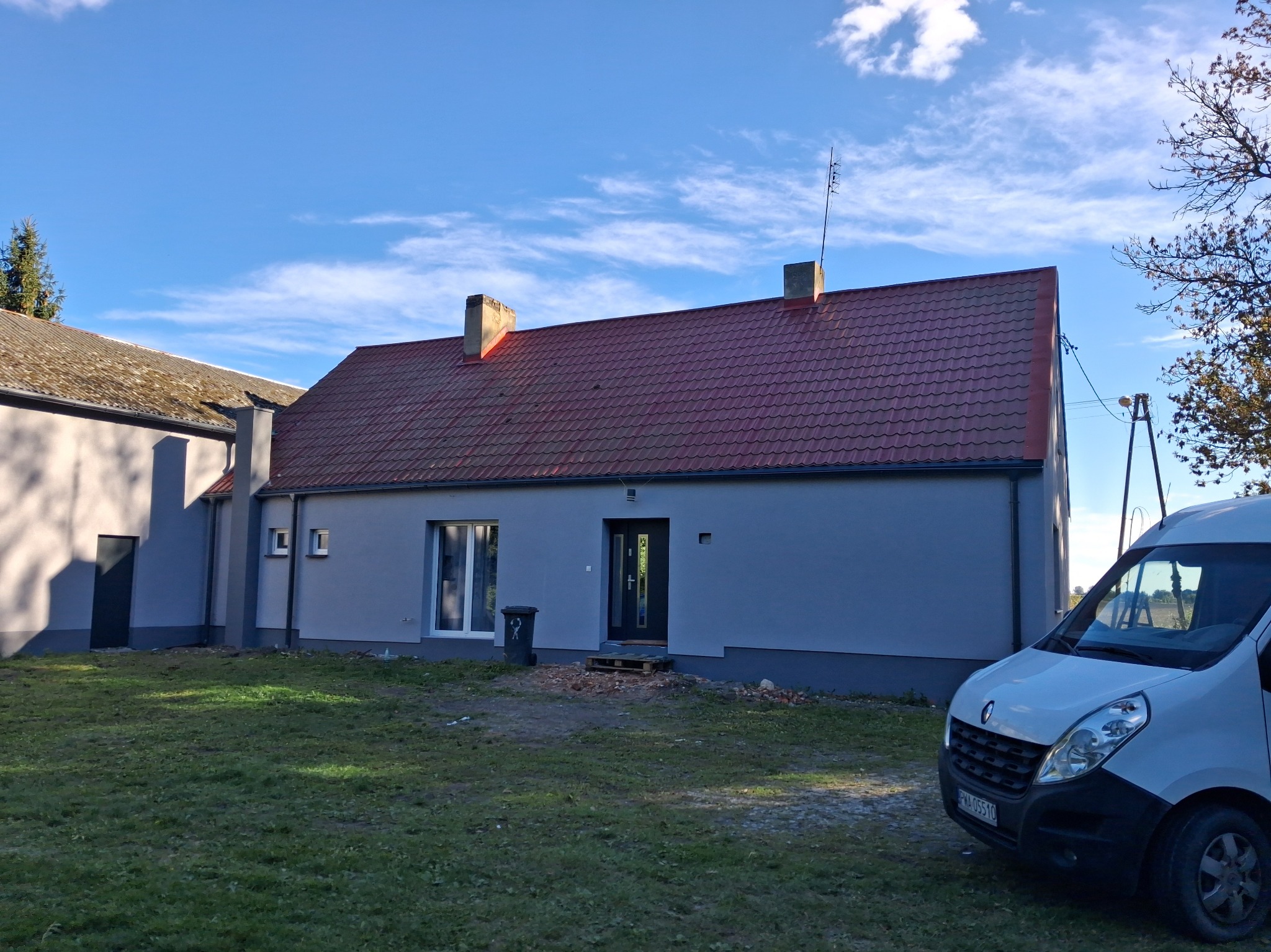 Odnowiony dom jednorodzinny z czerwoną dachówką i szarą elewacją, widoczny z zewnątrz, z samochodem dostawczym w tle. Świeżo pomalowane ściany i nowe okna.