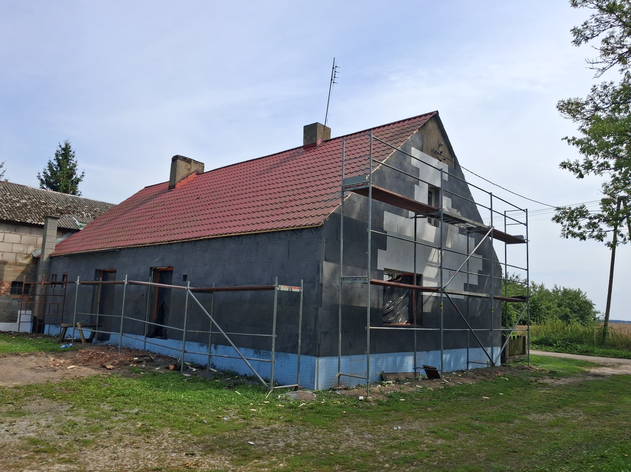 Dom w trakcie ocieplania, widoczne rusztowania i czarny materiał izolacyjny na ścianach. Czerwony dach i zielone otoczenie. Etap prac w słoneczny dzień.