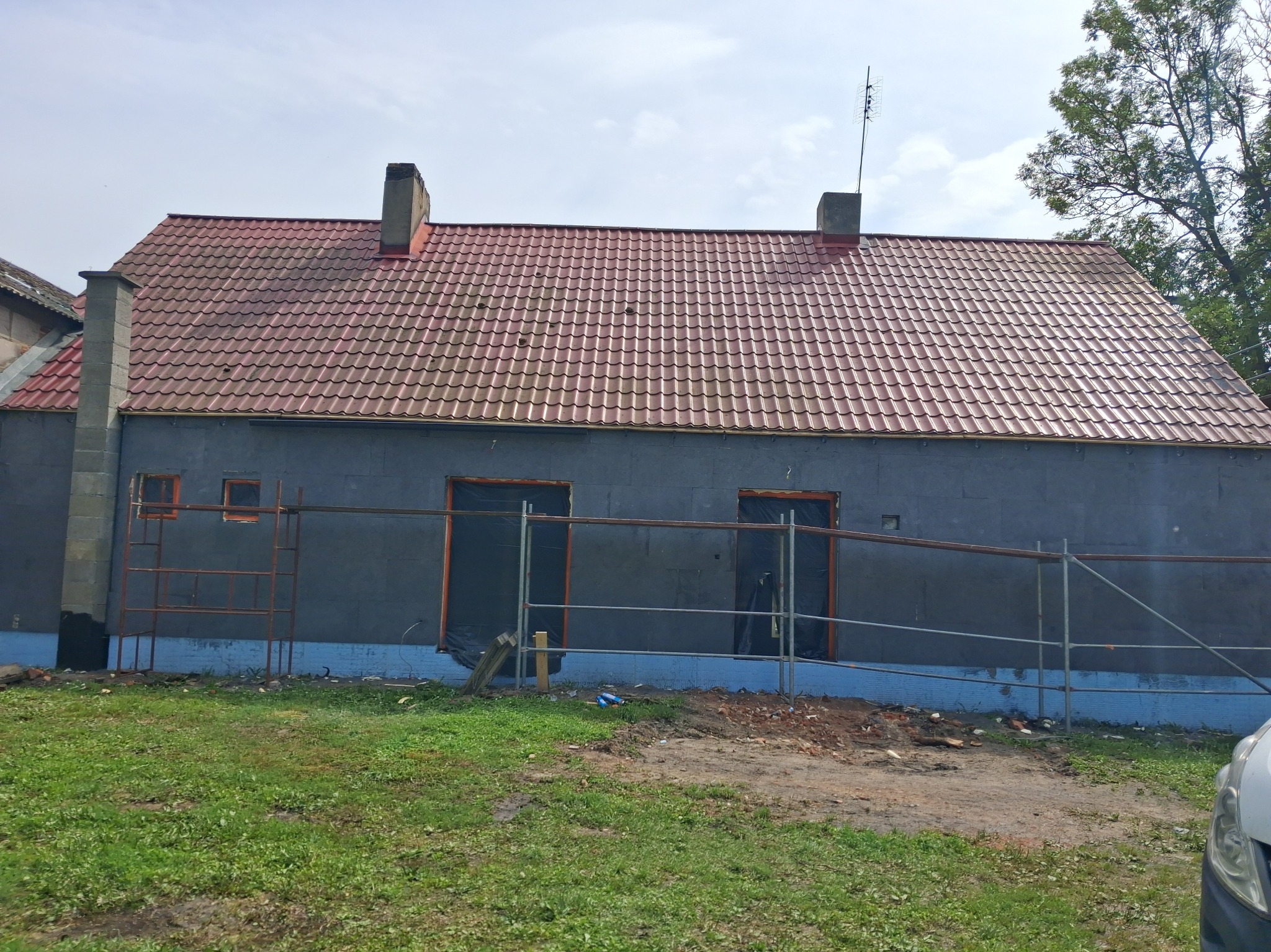 Ocieplony budynek z czerwonym dachem, widoczne rusztowania i okna zabezpieczone folią. Prace w toku, widok z poziomu trawnika.