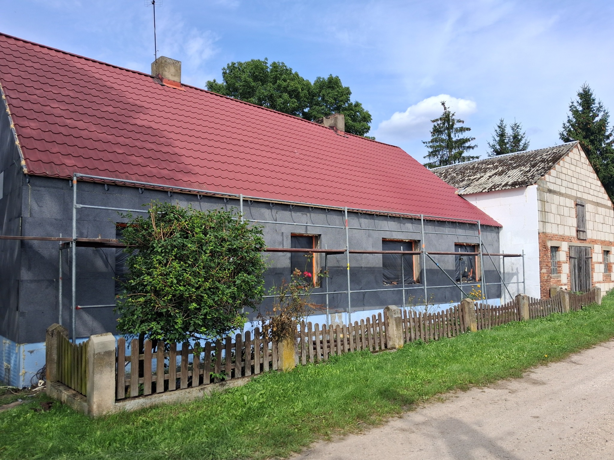Dom w trakcie ocieplania: szara elewacja zabezpieczona siatką, czerwony dach, rusztowania. Widoczny fragment drewnianego płotu i trawnika. Ujęcie z poziomu gruntu.