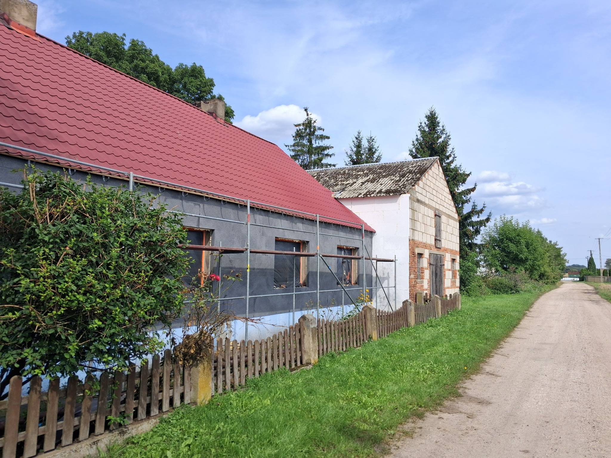 Dom w trakcie ocieplania, widoczne rusztowania i materiał izolacyjny na ścianach. Czerwony dach i drewniany płot wzdłuż drogi gruntowej. Częściowy widok na sąsiedni budynek.