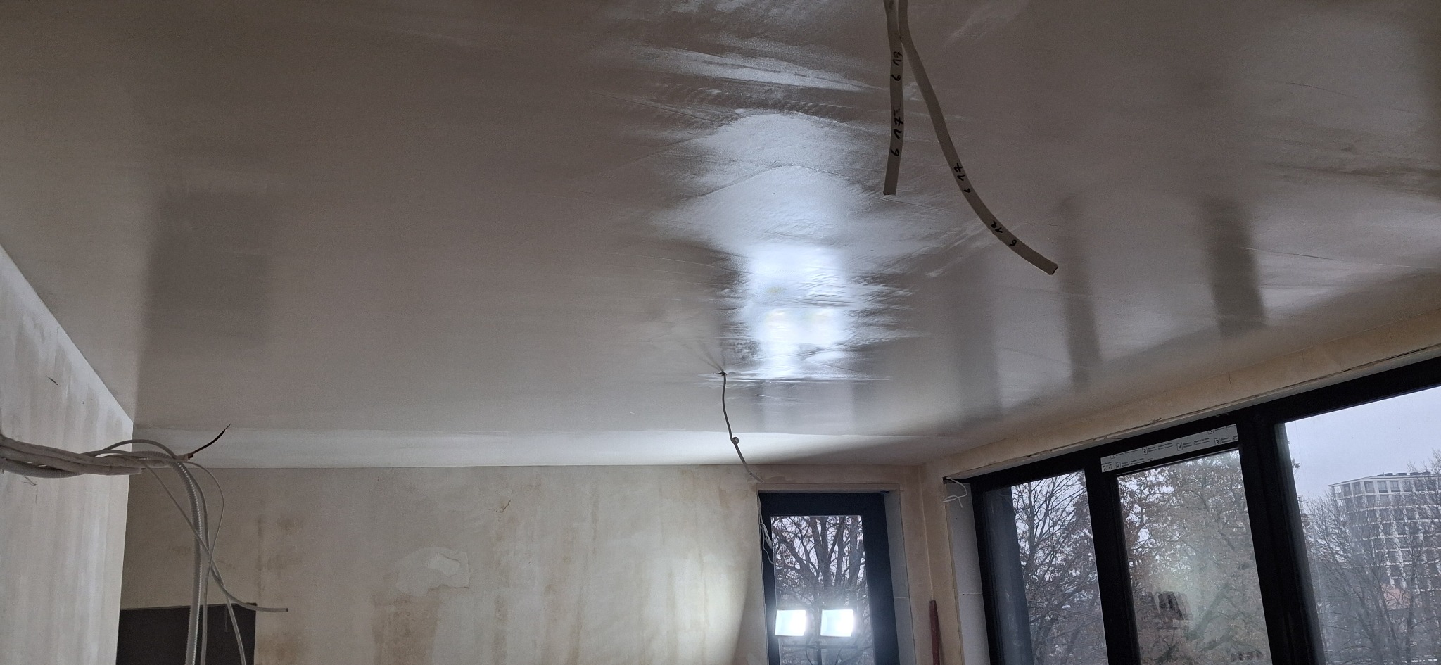 Gładko wyszpachlowany sufit w trakcie remontu mieszkania, z widocznymi przewodami elektrycznymi i oknem z widokiem na budynek. Świeżo nałożona gładź szpachlowa.