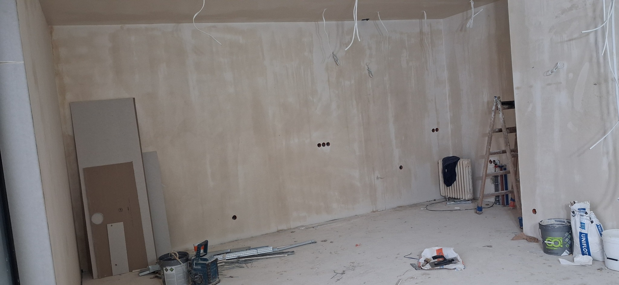 Pokój w trakcie szpachlowania ścian, widoczne płyty karton-gips, drabina, puszki elektryczne i materiały budowlane. Ściany w jasnym odcieniu beżu, surowe wykończenie.