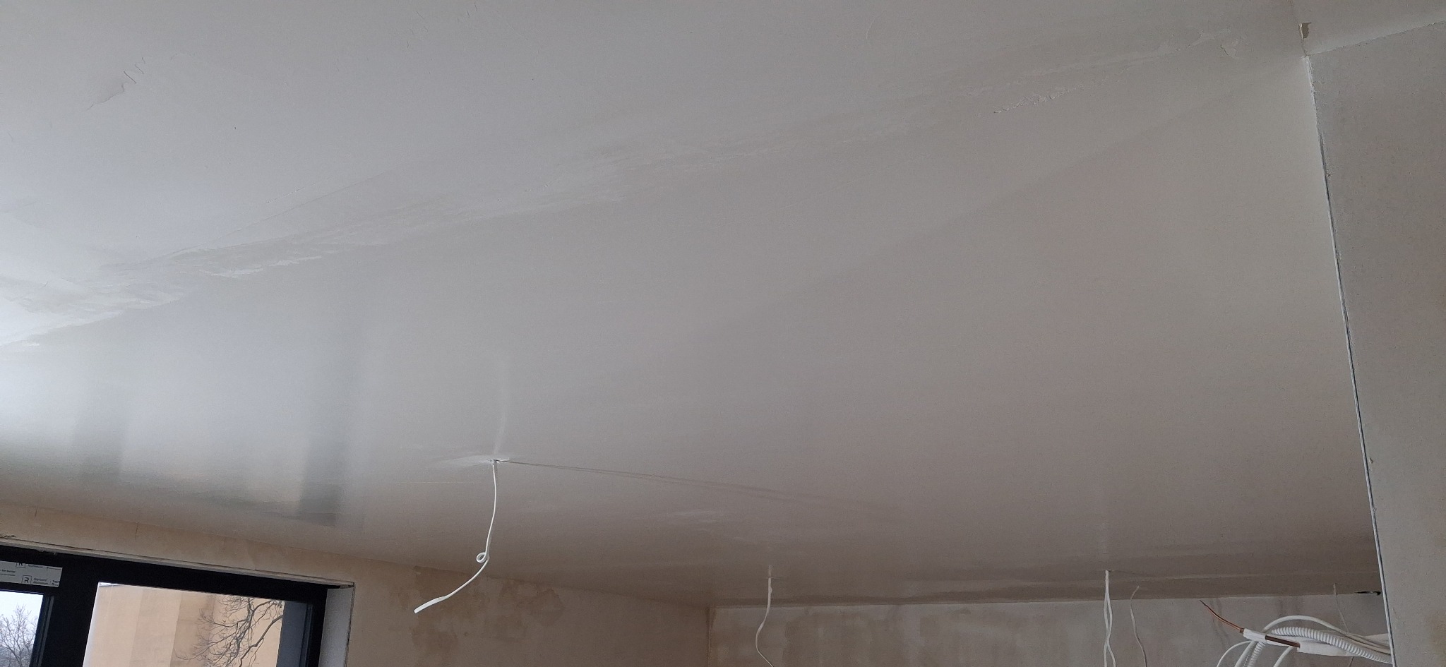 Gładka, biała powierzchnia sufitu po szpachlowaniu, widoczne przewody elektryczne. Fragment okna w lewym dolnym rogu. Świeżo wykończone wnętrze w trakcie prac.