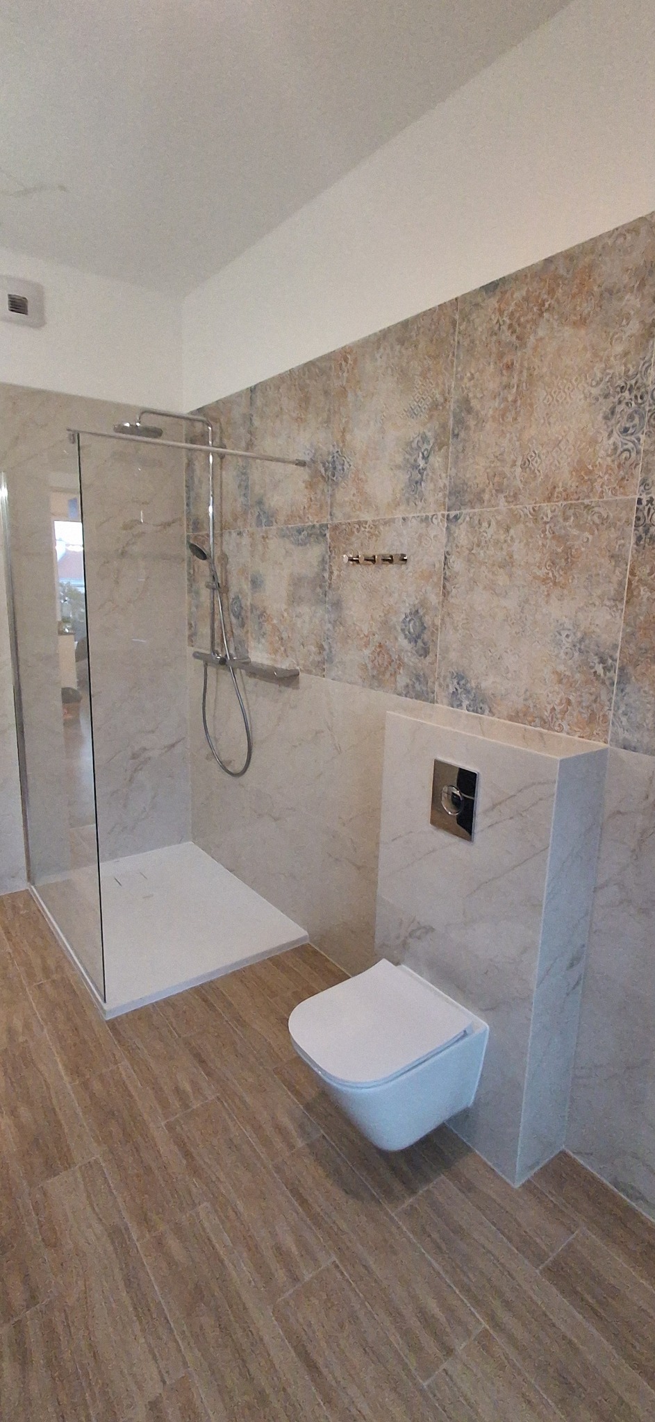 Nowoczesna łazienka po remoncie: kabina prysznicowa typu walk-in, podwieszana toaleta i płytki imitujące drewno na podłodze. Ściany wykończone płytkami w stylu patchwork.