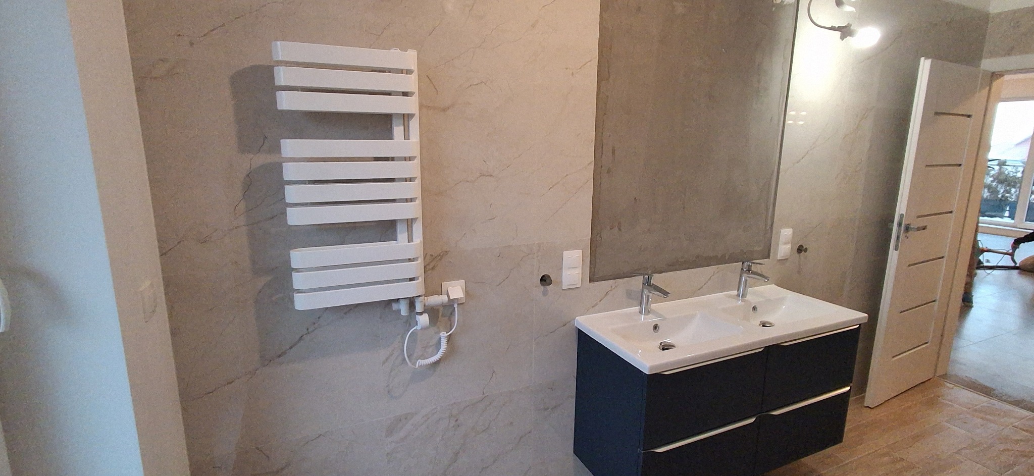 Wykończona łazienka z grzejnikiem drabinkowym, umywalką z szafką i dużym lustrem. Ściany wyłożone płytkami imitującymi marmur. Widoczny fragment otwartych drzwi.