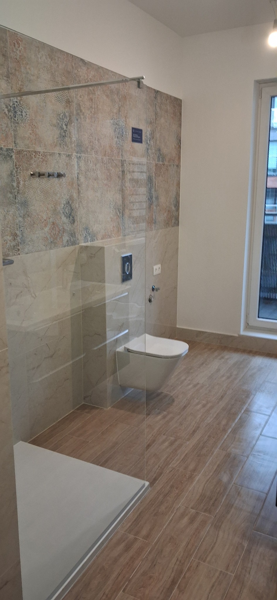 Nowoczesna łazienka z kabiną prysznicową typu walk-in, wiszącą toaletą i płytkami imitującymi drewno. Ściany wykończone dekoracyjnymi płytkami w odcieniach beżu i brązu.