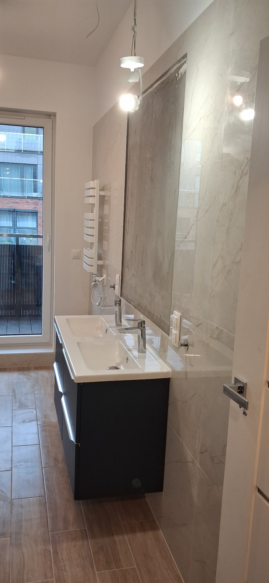 Wykończona łazienka z podwójną umywalką, grzejnikiem drabinkowym i marmurowymi płytkami. Widok na balkon przez okno. Nowoczesne oświetlenie sufitowe.